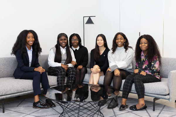 Six female Urban Synergy Mentee graduates sit on a grey corner sofa in an office. All are dressed smartly in suits or office clothing, and smiling at the camera.