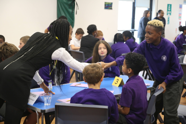 A school seminar. Students sit around a blue table wearing a purple uniform. Leila leans over the desk to shake the hand of a young boy who is also standing at the opposite end of the table. Other groups can be seen in the background sat at tables and talking amongst themselves.